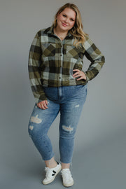 A woman with blonde hair stands against a plain gray background, smiling with one hand on her hip. She wears the Olive & Black Plaid Button Up from the Panache Apparel Collection a super soft, long-sleeve shirt that adds comfort to her casual look paired with blue ripped jeans and white sneakers.