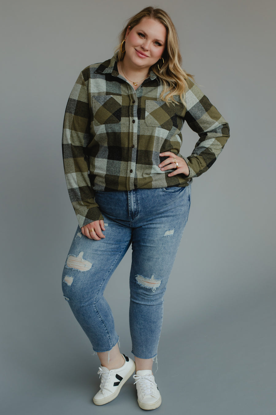 A woman with blonde hair stands against a plain gray background, smiling with one hand on her hip. She wears the Olive & Black Plaid Button Up from the Panache Apparel Collection a super soft, long-sleeve shirt that adds comfort to her casual look paired with blue ripped jeans and white sneakers.