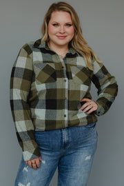 A person wearing the Panache Apparel Collection Olive & Black Plaid Button Up and distressed blue jeans stands against a plain gray background, smiling with one hand on their hip.