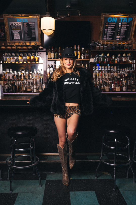 A woman wearing a cowboy hat, black fur coat, Panache Apparel Collection Black Whiskey Weather Tank Top, leopard print shorts, and boots stands in a bar with shelves of liquor bottles and draft menus in the background.