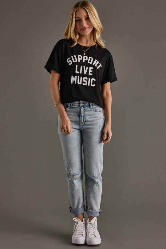 A woman smiles against a plain background, wearing the Panache Apparel Collection's Support Live Music Tee, light blue ripped jeans, white sneakers, and gold jewelry.