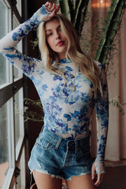A woman with long blonde hair models the Panache Apparel Collection Blue & White Floral Mesh Top and denim shorts indoors by a large window and green plant, raising one arm above her head.
