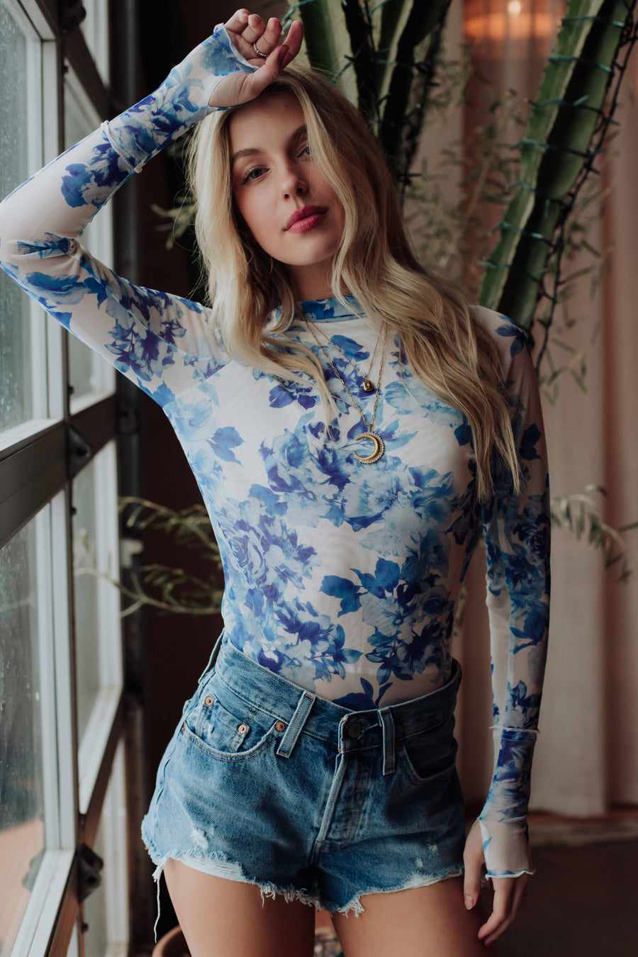 A woman with long blonde hair models the Panache Apparel Collection Blue & White Floral Mesh Top and denim shorts indoors by a large window and green plant, raising one arm above her head.