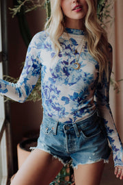 A woman wears the Panache Apparel Collection Blue & White Floral Mesh Top, layered gold necklaces, and high-waisted denim shorts. She stands indoors by a window and potted plant, her face partially out of frame.