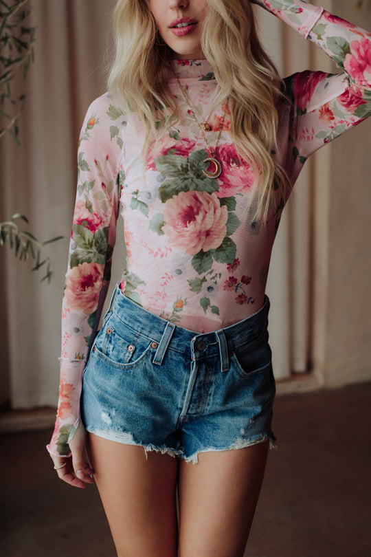 A woman with long blonde hair wears the Panache Apparel Collection Pink & Green Floral Mesh Top with high-waisted denim shorts and layered gold necklaces. Her face is partially out of frame.