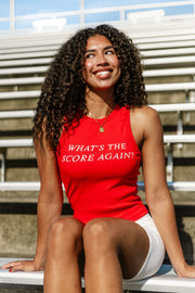A smiling woman with curly hair sits on bleachers wearing the Panache Apparel Collection Red What's The Score Again? Tank Top, featuring a ribbed design and crew neckline, paired with white shorts.