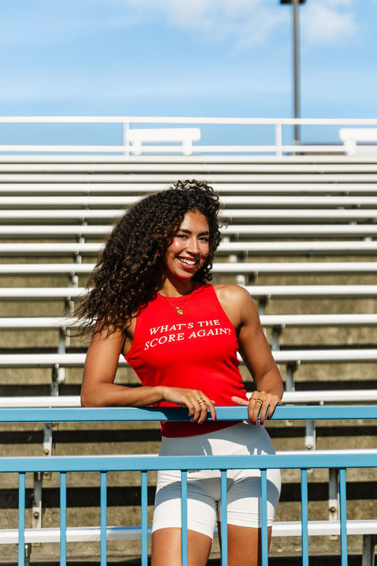 A woman with curly hair stands and smiles in front of empty stadium bleachers, holding a blue railing, wearing the Panache Apparel Collection Red What's The Score Again? Tank Top.
