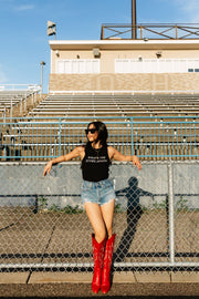 Wearing the Panache Apparel Collection's black What's The Score Again? Tank Top, denim shorts, red cowboy boots, and sunglasses, a woman leans against a chain-link fence in front of empty stadium bleachers on a sunny day, as if pondering the score.