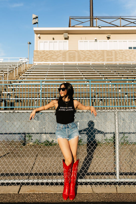 Wearing the Panache Apparel Collection's black What's The Score Again? Tank Top, denim shorts, red cowboy boots, and sunglasses, a woman leans against a chain-link fence in front of empty stadium bleachers on a sunny day, as if pondering the score.