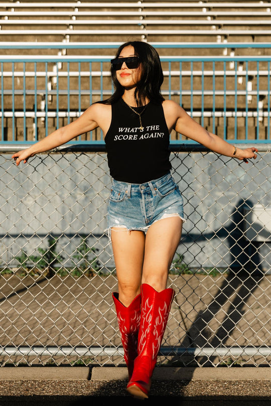 A woman in sunglasses, denim shorts, and red cowboy boots leans against a chain-link fence in front of empty bleachers, wearing the Black What's The Score Again? Tank Top from Panache Apparel Collection.