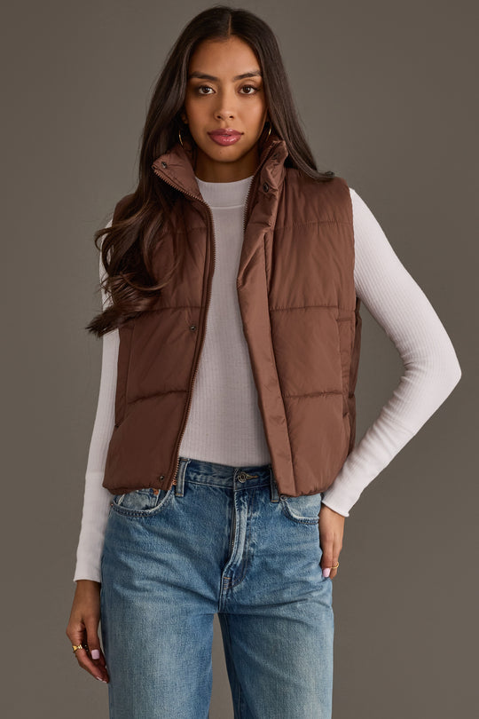 A woman models the Panache Apparel Collection’s Brown Vest, layered over a white long-sleeve shirt and blue jeans, as she stands against a plain gray background and gazes at the camera with a neutral expression.