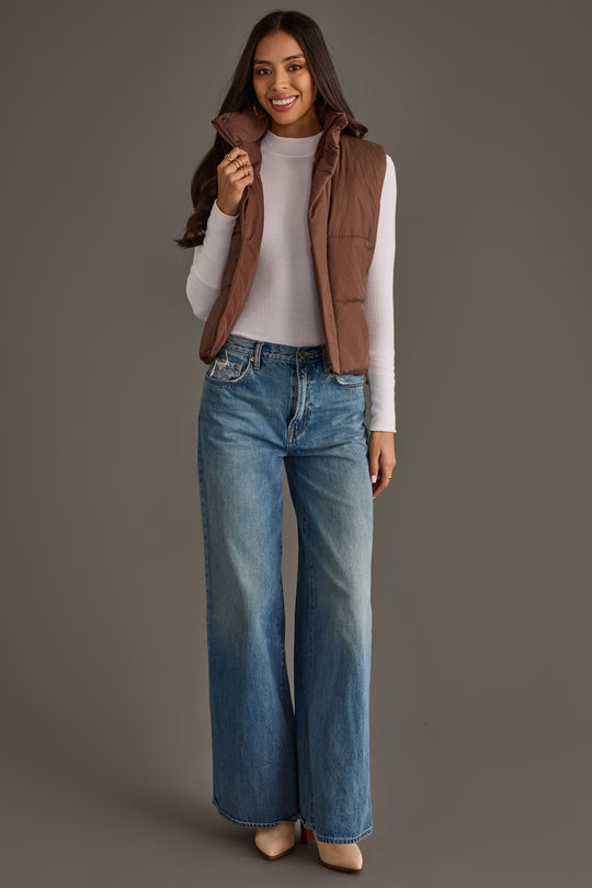 A woman stands smiling against a plain gray background, wearing the Panache Apparel Collection Brown Vest over a white long-sleeve top, paired with wide-leg blue jeans and beige shoes.
