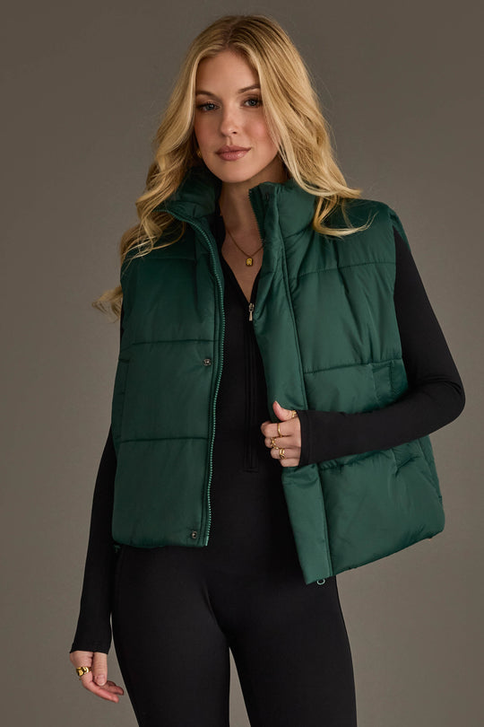 A woman with long blonde hair models the Panache Apparel Collection Dark Green Vest, layered over a black long-sleeve outfit, as she stands against a plain gray background.
