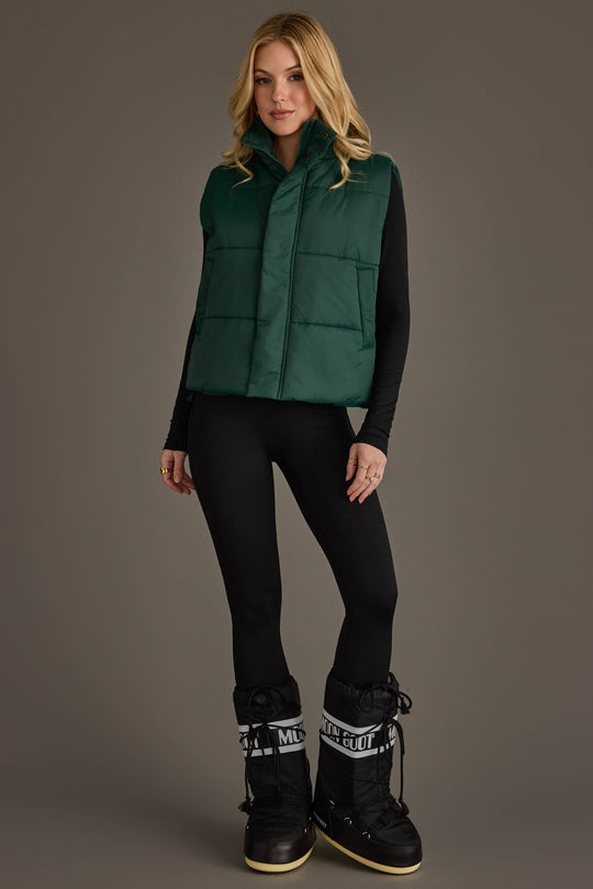 A woman with long blonde hair stands against a plain background wearing the Panache Apparel Collection Dark Green Vest over a black long-sleeve top, paired with black leggings and black snow boots. Her hands rest by her sides.