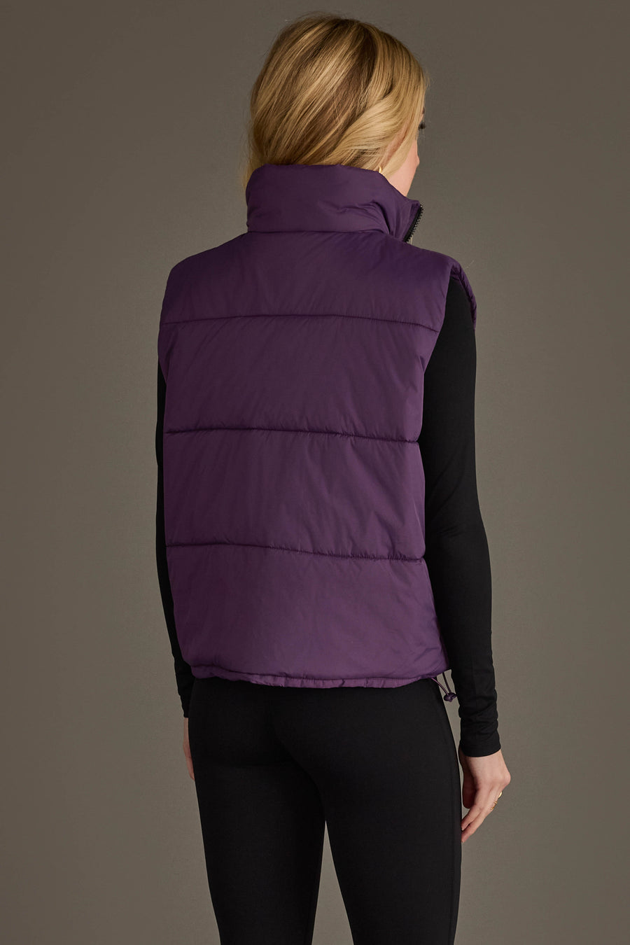 A person with long blonde hair stands with their back to the camera against a plain background, wearing the Panache Apparel Collection’s Dark Purple Vest over a black long-sleeve top and black leggings.