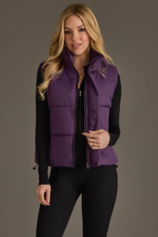 A woman with long blonde hair models the Panache Apparel Collection Dark Purple Vest over a black long-sleeve top and black pants, standing against a plain gray background.