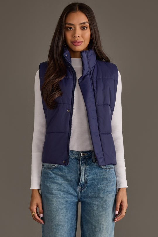 A woman models the Panache Apparel Collection Navy Vest, layered over a white long-sleeve top and paired with blue jeans, standing against a plain gray background.