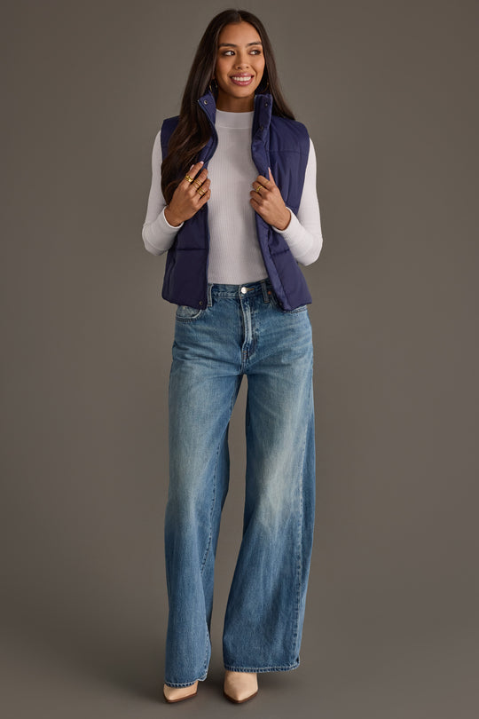 A woman stands smiling against a plain background, wearing the Panache Apparel Collection Navy Vest over a white long-sleeve top, paired with blue wide-leg jeans and beige pointed-toe shoes.