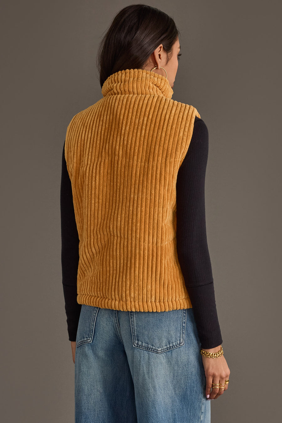 A woman with long dark hair is seen from behind, wearing the Panache Apparel Collection’s Mustard Corduroy Vest over a black long-sleeve shirt and blue jeans, set against a plain gray background.