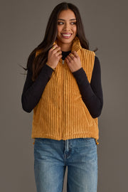 A woman smiles while holding the collar of the Panache Apparel Collection Mustard Corduroy Vest, styled over a black long-sleeve shirt and blue jeans, against a plain neutral background.