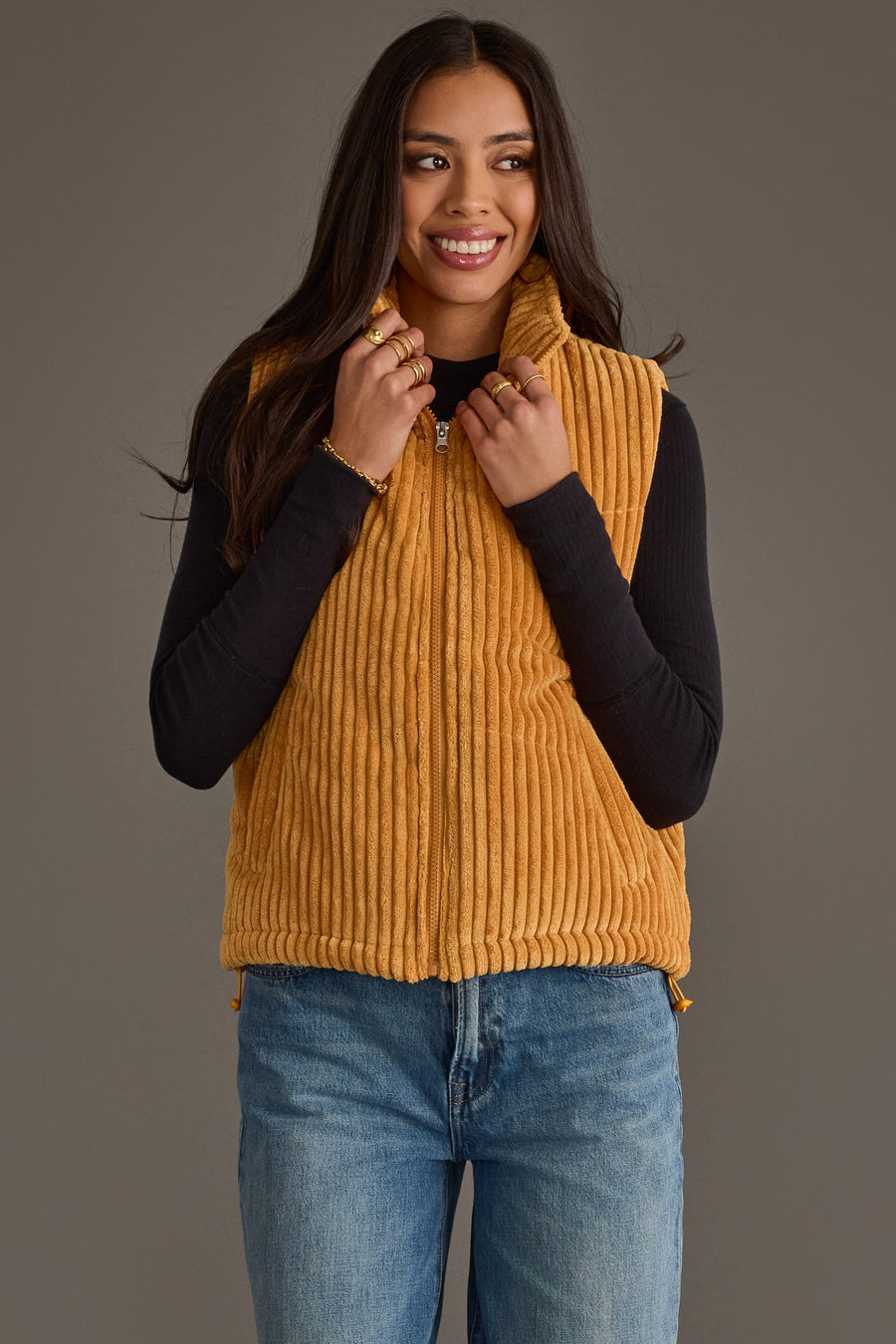 A woman smiles while holding the collar of the Panache Apparel Collection Mustard Corduroy Vest, styled over a black long-sleeve shirt and blue jeans, against a plain neutral background.