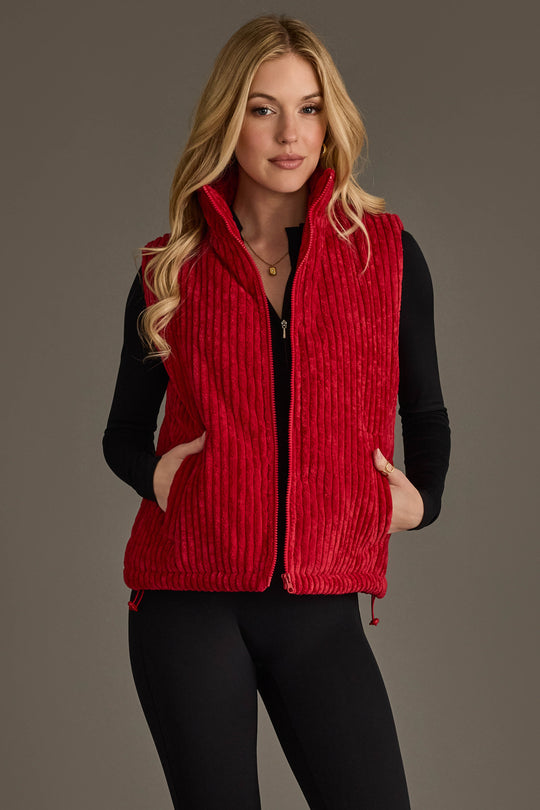 A woman with long blonde hair stands against a plain gray background, wearing the Panache Apparel Collection’s Dark Red Corduroy Vest over a black long-sleeve top and black pants, her hands tucked into the vest’s pockets.