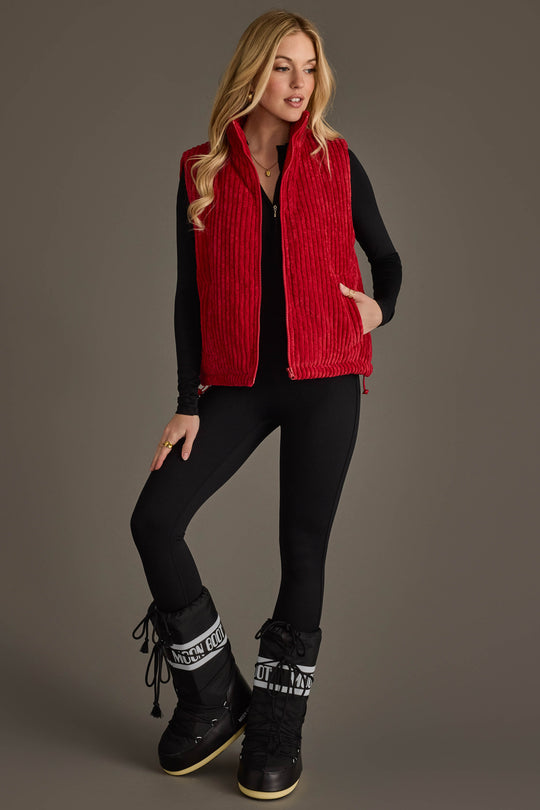 A woman stands against a plain brown background with one hand in her pocket, wearing the Panache Apparel Collection Dark Red Corduroy Vest over a black long-sleeve top and black pants, paired with black snow boots featuring white accents.