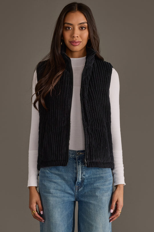 A woman faces forward against a plain gray background, wearing the Panache Apparel Collection Black Corduroy Vest layered over a white ribbed long-sleeve top and blue jeans. Her long hair is down and her hands rest relaxed at her sides.