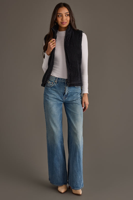 A woman with long dark hair stands against a gray background, smiling softly with one hand near her collar. She is wearing the Panache Apparel Collection Black Corduroy Vest over a white long-sleeve top, paired with wide-leg blue jeans and beige shoes.