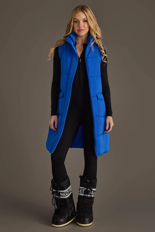 A woman models the Panache Apparel Collection’s Royal Blue Long Vest over a black outfit and black snow boots, standing against a plain background and looking at the camera with a relaxed expression.