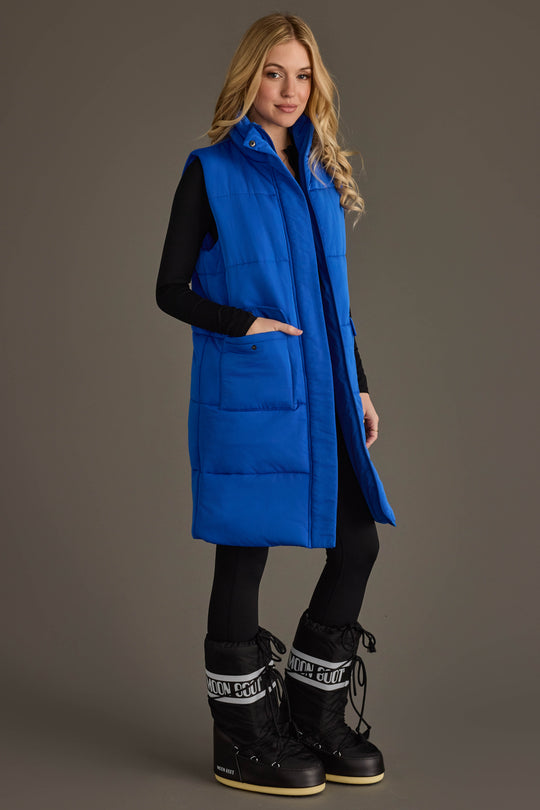 A person wearing the Panache Apparel Collection’s Royal Blue Long Vest over a black outfit and black snow boots stands against a plain gray background, looking at the camera and smiling slightly.