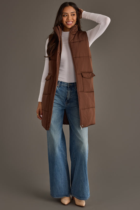 A woman wearing the Panache Apparel Collection’s Dark Brown Long Vest over a white long-sleeve top and flared blue jeans smiles with one hand on her head, standing against a plain gray background.