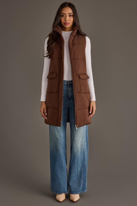 A woman models the Panache Apparel Collection Dark Brown Long Vest, styled over a white sweater with wide-leg blue jeans and beige shoes, standing facing forward against a plain gray background.