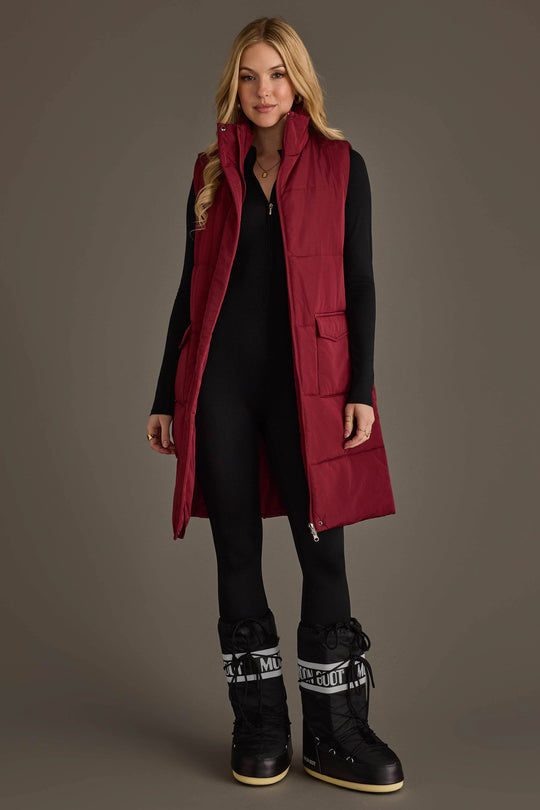 A woman stands against a plain background, wearing the Burgundy Long Vest from Panache Apparel Collection over a black jumpsuit and black snow boots featuring white MOON BOOT text on the cuffs.