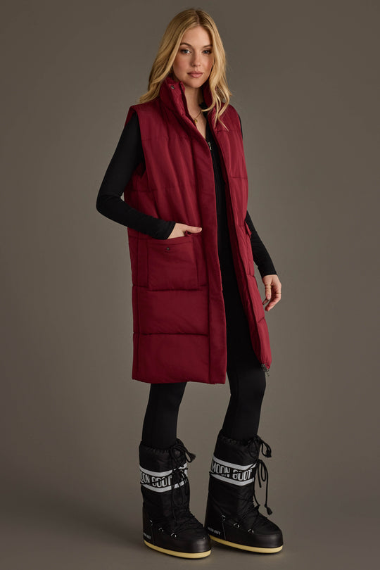 A woman wearing the Panache Apparel Collection Burgundy Long Vest, paired with a black long-sleeve top, black leggings, and black snow boots, stands against a plain gray background.