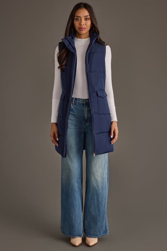 A woman stands against a gray background, modeling the Panache Apparel Collection Navy Long Vest layered over a white turtleneck sweater, paired with wide-leg blue jeans and beige heels, her hands resting relaxed at her sides.