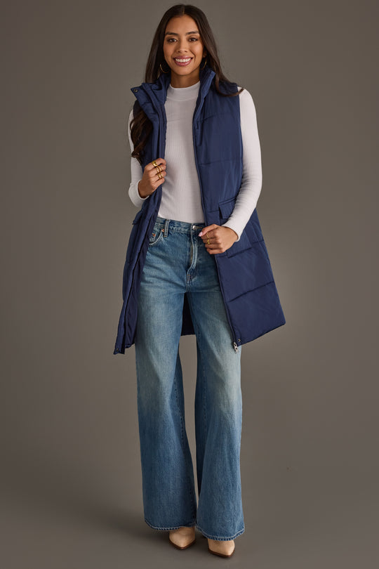 A woman stands and smiles against a plain gray background, wearing the Panache Apparel Collection Navy Long Vest over a white turtleneck, paired with wide-leg jeans and beige heels.