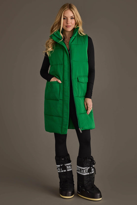 A woman models the Panache Apparel Collection’s Classic Green Long Vest over a black outfit and black snow boots, standing against a plain background with one hand in her vest pocket and the other resting at her side.
