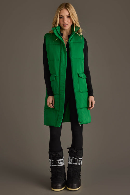 A person stands facing forward against a plain brown background, wearing the Panache Apparel Collection Classic Green Long Vest over a black outfit and black snow boots with white details.