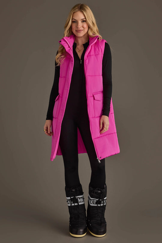 A woman with light brown wavy hair stands facing forward and smiling against a plain gray background, wearing the Panache Apparel Collection’s Fuchsia Long Vest over a black outfit and black winter boots.