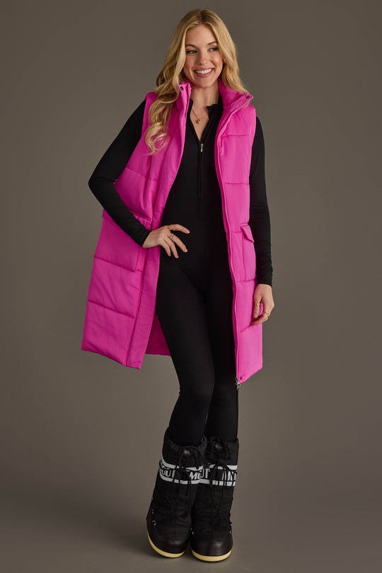 A woman stands smiling against a plain gray background, wearing the Panache Apparel Collection’s Fuchsia Long Vest over a black outfit and black snow boots.