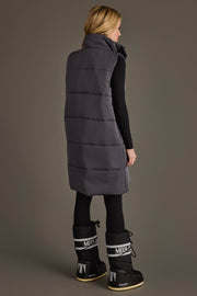 A woman stands against a plain gray background, facing away and wearing the Panache Apparel Collection Dark Gray Long Vest layered over black leggings and top, paired with black and white Moon Boot winter boots.