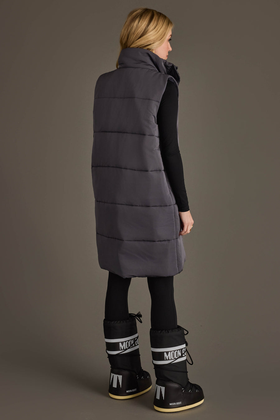 A woman stands against a plain gray background, facing away and wearing the Panache Apparel Collection Dark Gray Long Vest layered over black leggings and top, paired with black and white Moon Boot winter boots.