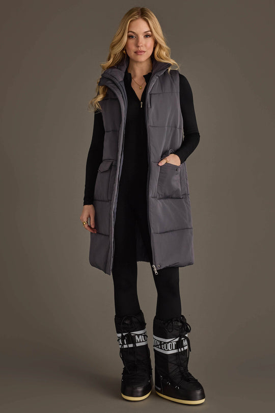 A woman stands facing forward wearing the Panache Apparel Collection Dark Gray Long Vest over a black fitted outfit and black snow boots with white lettering. She has long blonde hair, a neutral expression, and her hands are in the vest pockets.