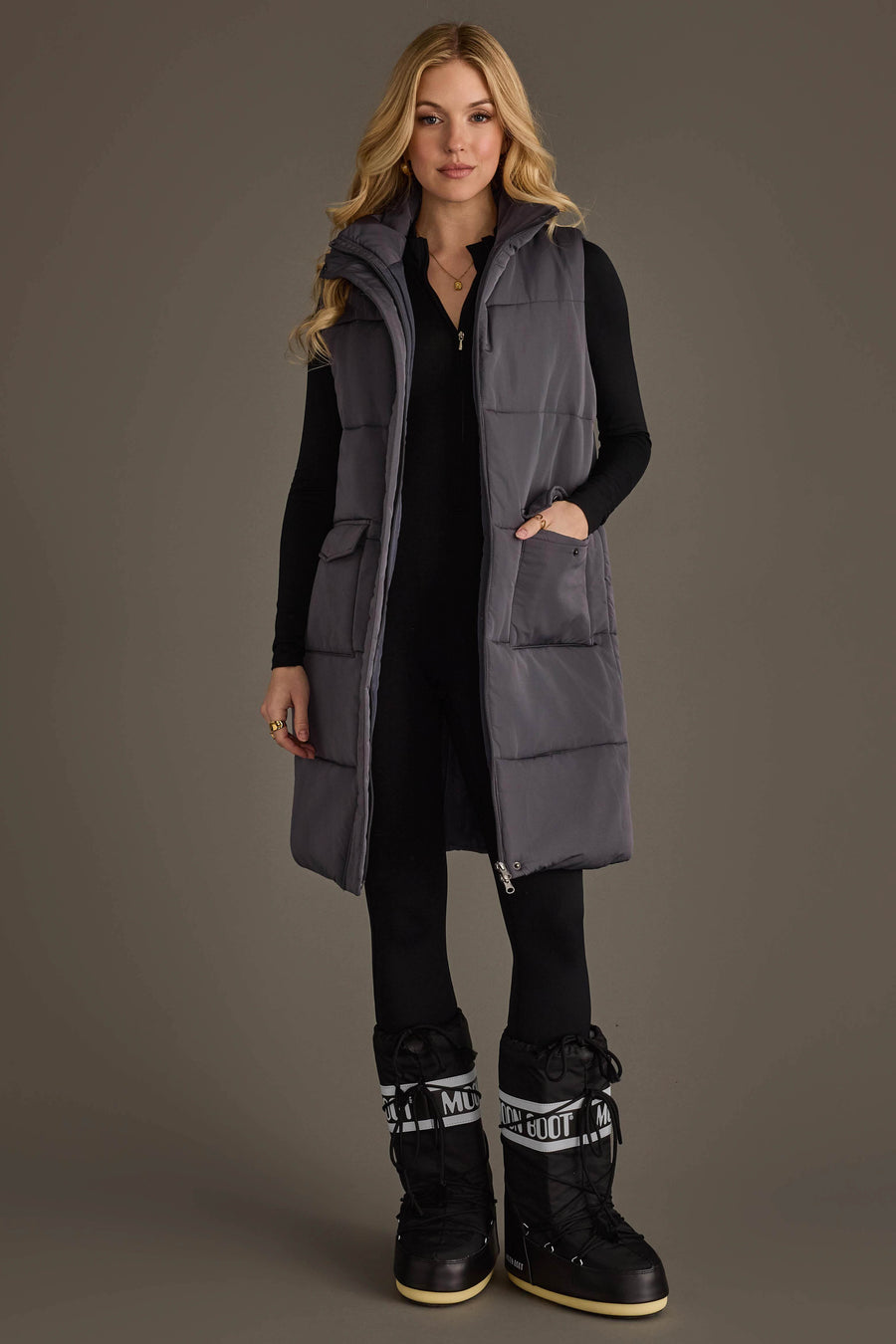 A woman stands facing forward wearing the Panache Apparel Collection Dark Gray Long Vest over a black fitted outfit and black snow boots with white lettering. She has long blonde hair, a neutral expression, and her hands are in the vest pockets.