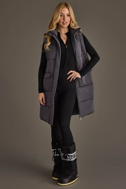 A woman with long blonde hair smiles while standing against a plain gray background, wearing a black jumpsuit and black snow boots with white laces, layered with the Dark Gray Long Vest from Panache Apparel Collection.