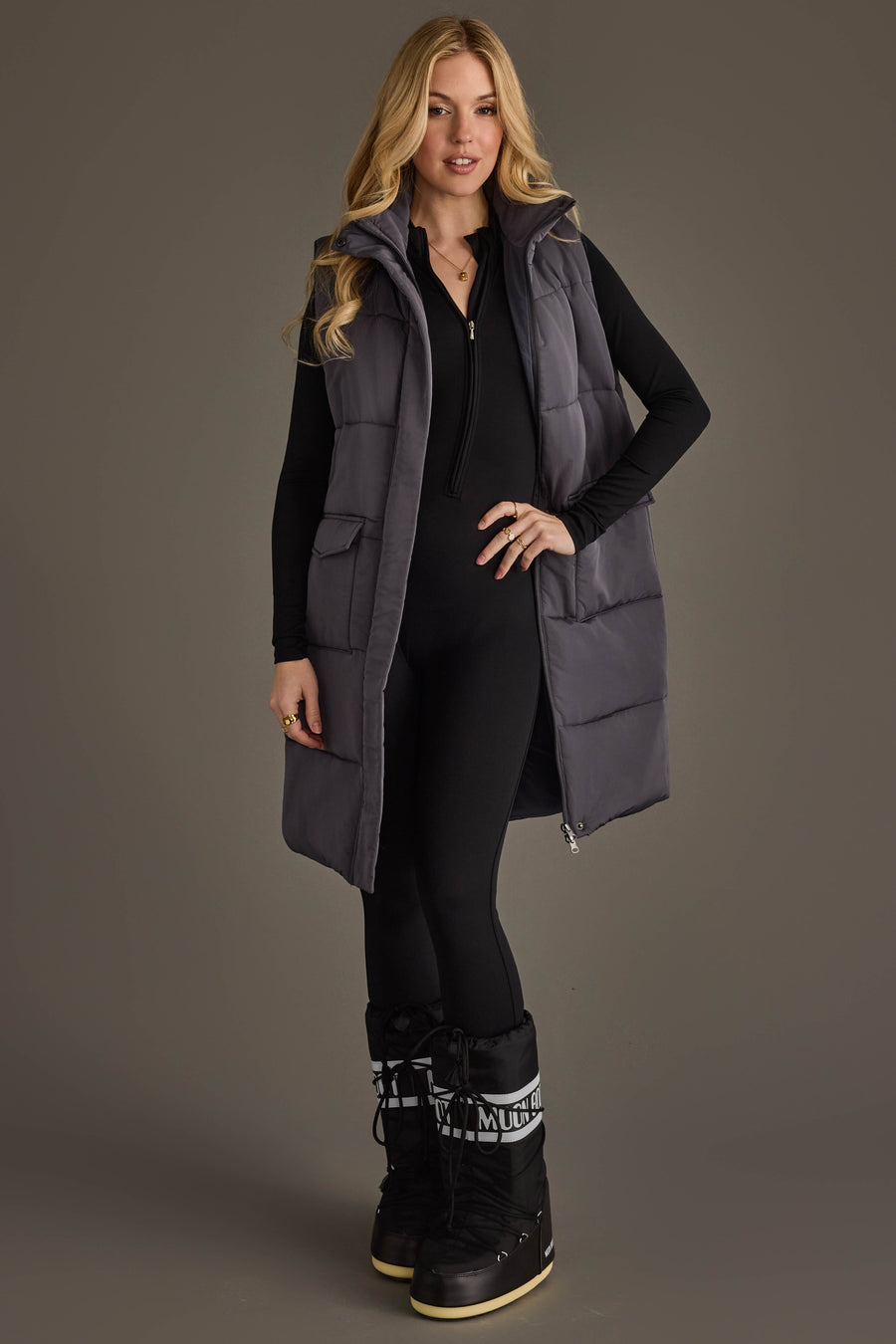 A woman with long blonde hair smiles while standing against a plain gray background, wearing a black jumpsuit and black snow boots with white laces, layered with the Dark Gray Long Vest from Panache Apparel Collection.