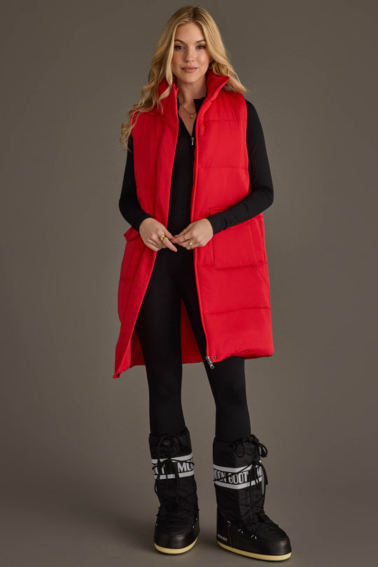 A blonde woman stands against a dark grey background, wearing the Red Long Vest from Panache Apparel Collection over a black long-sleeved top, black leggings, and black Moon Boot snow boots with white branding.