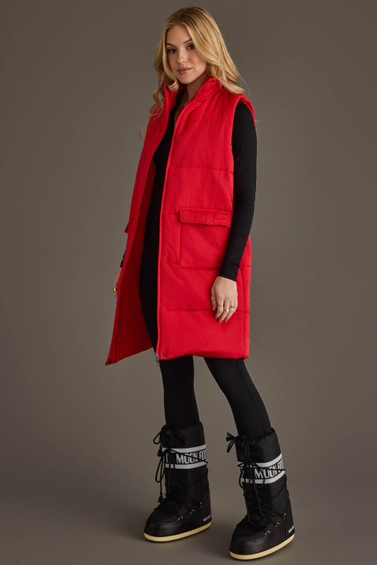 A woman models the Panache Apparel Collection Red Long Vest over a black outfit and black snow boots, standing and smiling against a plain gray background.