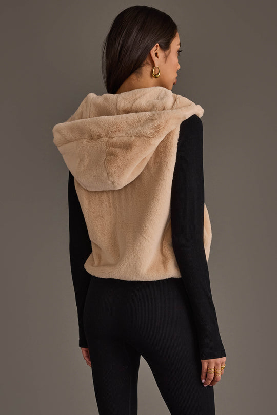 A woman with long dark hair is seen from the back wearing the Panache Apparel Collection Beige Faux Fur Vest over a black, long-sleeve ribbed outfit. She accessorizes with gold hoop earrings and rings.